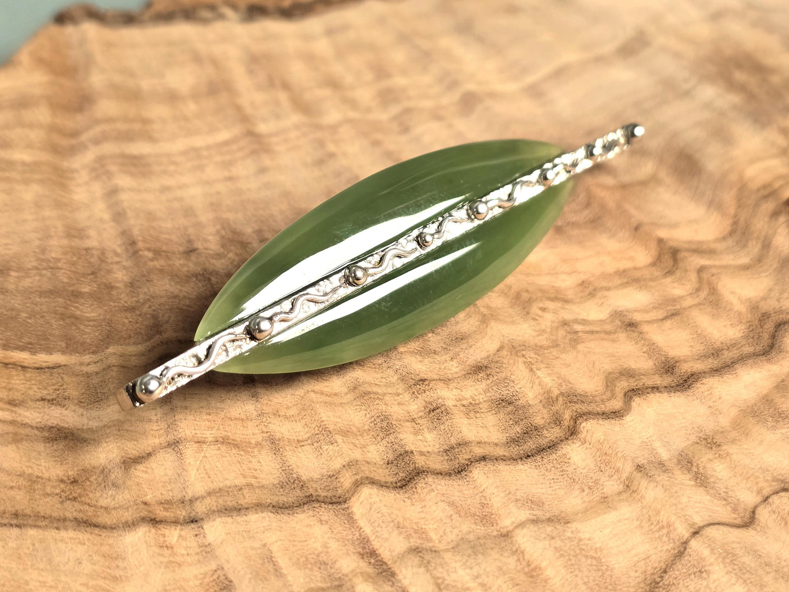 A green jade leaf-shaped brooch with a silver-toned bar, displayed on a textured wood slice background.