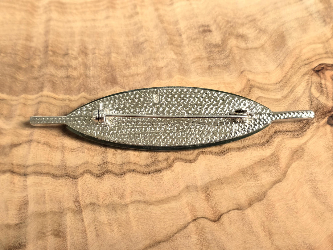 A silver-toned metal brooch shaped like a stylized leaf with a textured, woven pattern, displayed on a wavy wood grain background.