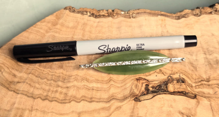 A black and white Sharpie fine liner marker placed on a natural wood slab next to a green leaf-shaped piece of silver glitter.