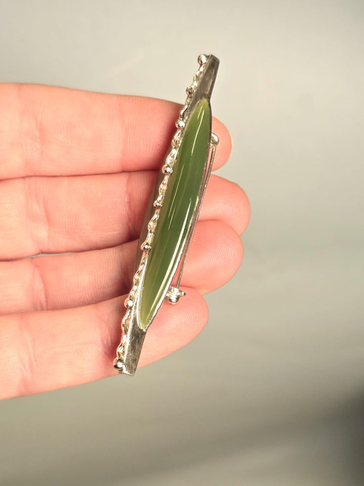 A silver bar brooch featuring a polished green jade inlay, held in a person&