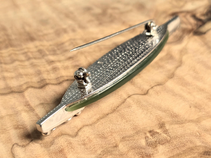 A silver-toned metal brooch with a textured handle and a green translucent inlay, resting on a wavy wood grain surface.