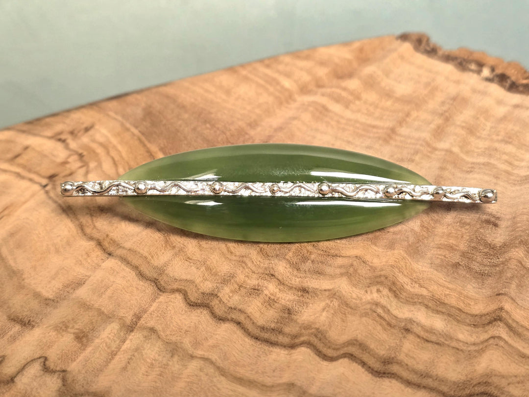 A green jade brooch with a silver-toned textured bar across the center, displayed on a wood slice background.