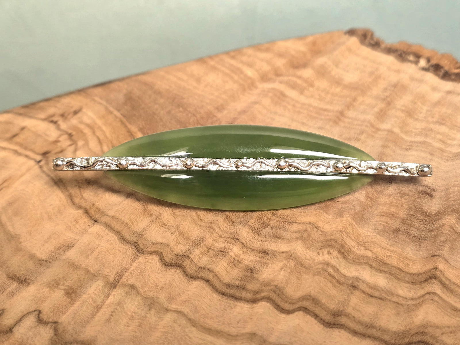 A green jade brooch with a silver-toned textured bar across the center, displayed on a wood slice background.