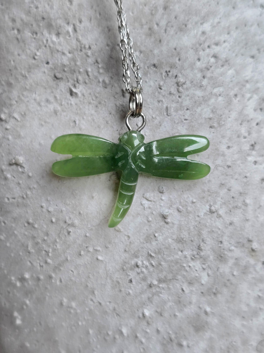 Green jade dragonfly pendant suspended from a chain against a textured light background