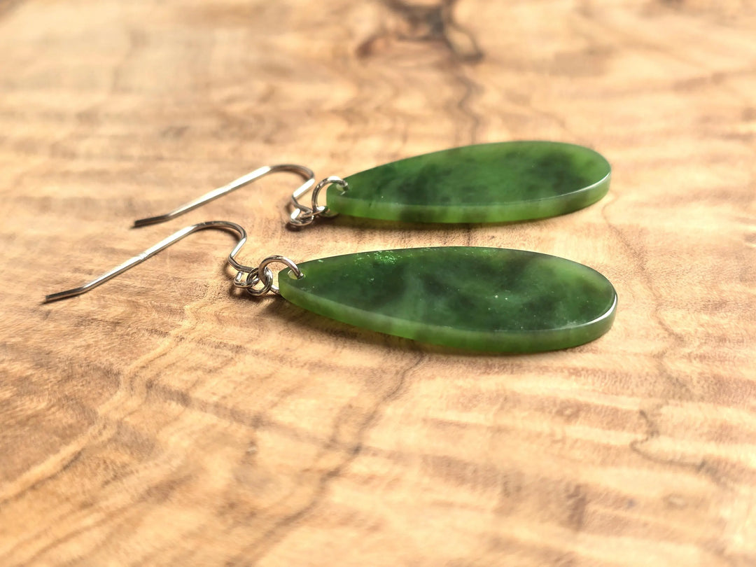 Pair of green jade teardrop earrings with silver hooks shown on wood in close up.