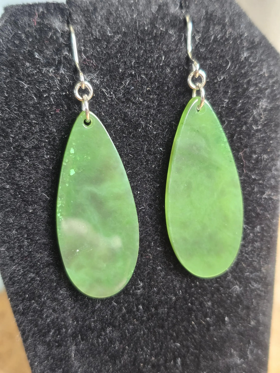 Two green jade teardrop earrings with silver hooks on a black display background.