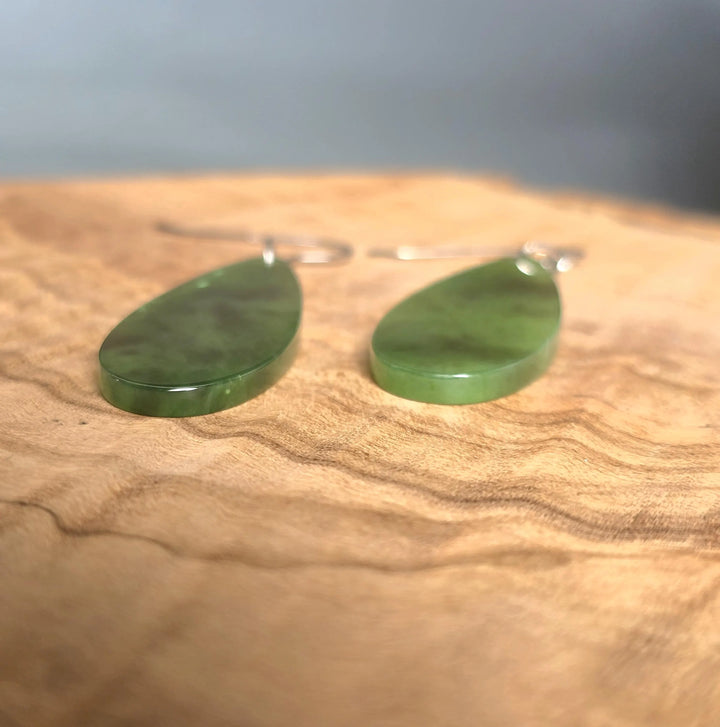 Green jade teardrop earrings with silver hooks resting on a wooden surface.