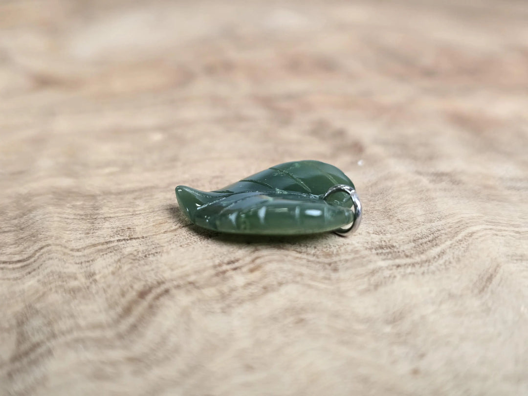 Green jade leaf pendant with a silver bail on a wooden surface.