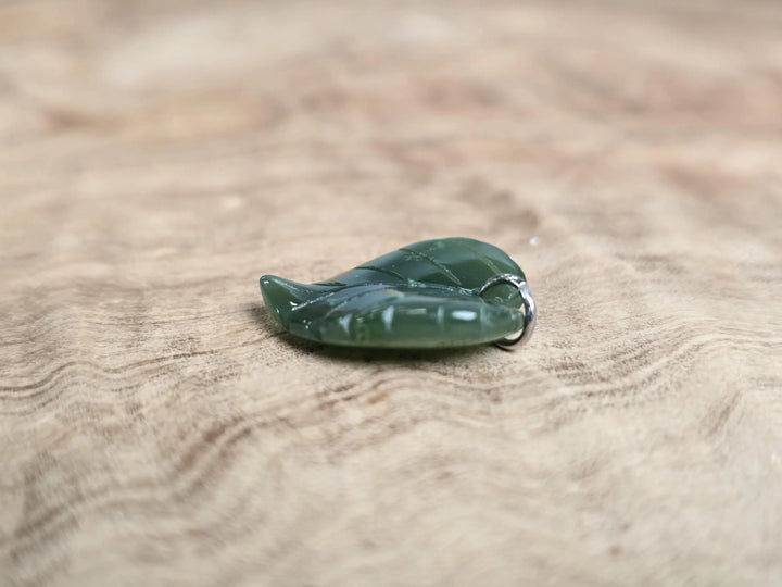 Green jade leaf pendant with a silver bail on a wooden surface.