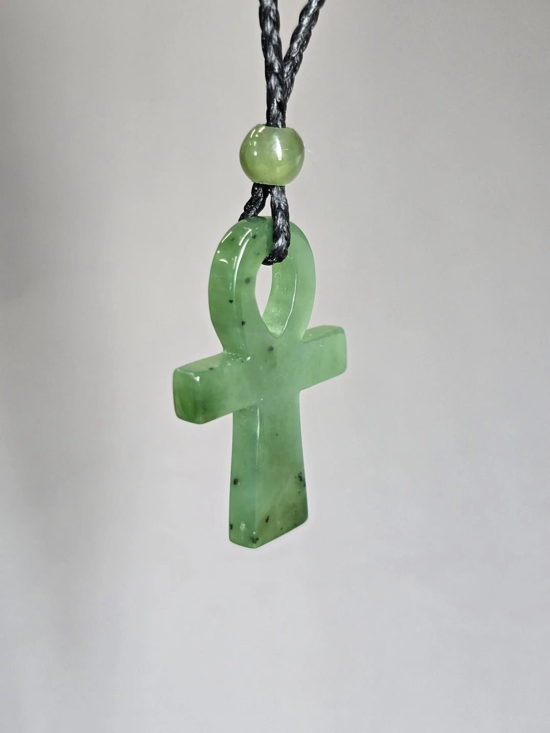 Nephrite jade ankh pendant in pale green on a black braided cord with bead near the top against wood background