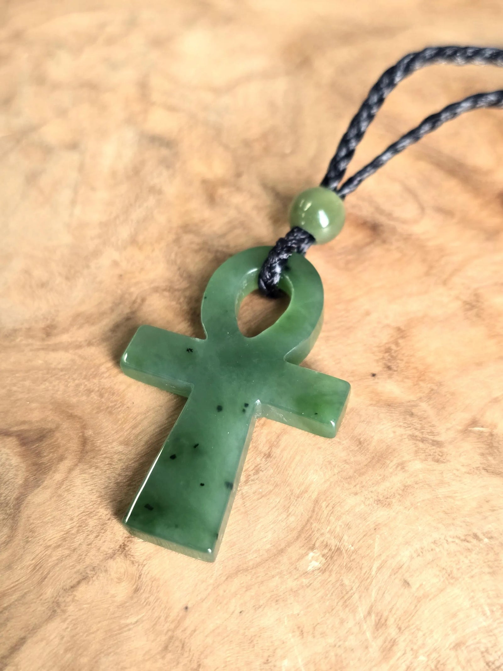 Close up of green nephrite jade ankh pendant on a black braided cord against a wooden surface