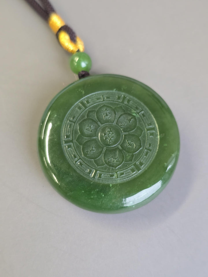 Green jade round pendant with intricate circular carving resting on a pale surface with a brown knot and bead.