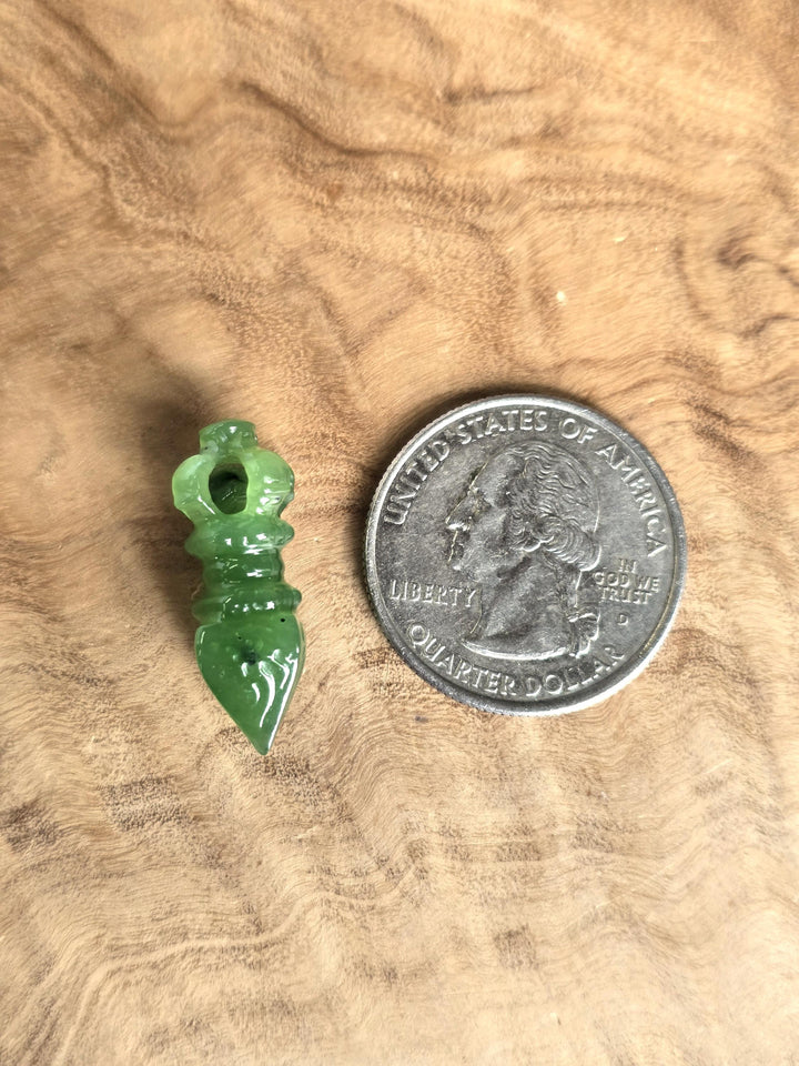 A small carved green stone pendant or charm shown next to a U.S. quarter coin for scale, placed on a textured wood surface.