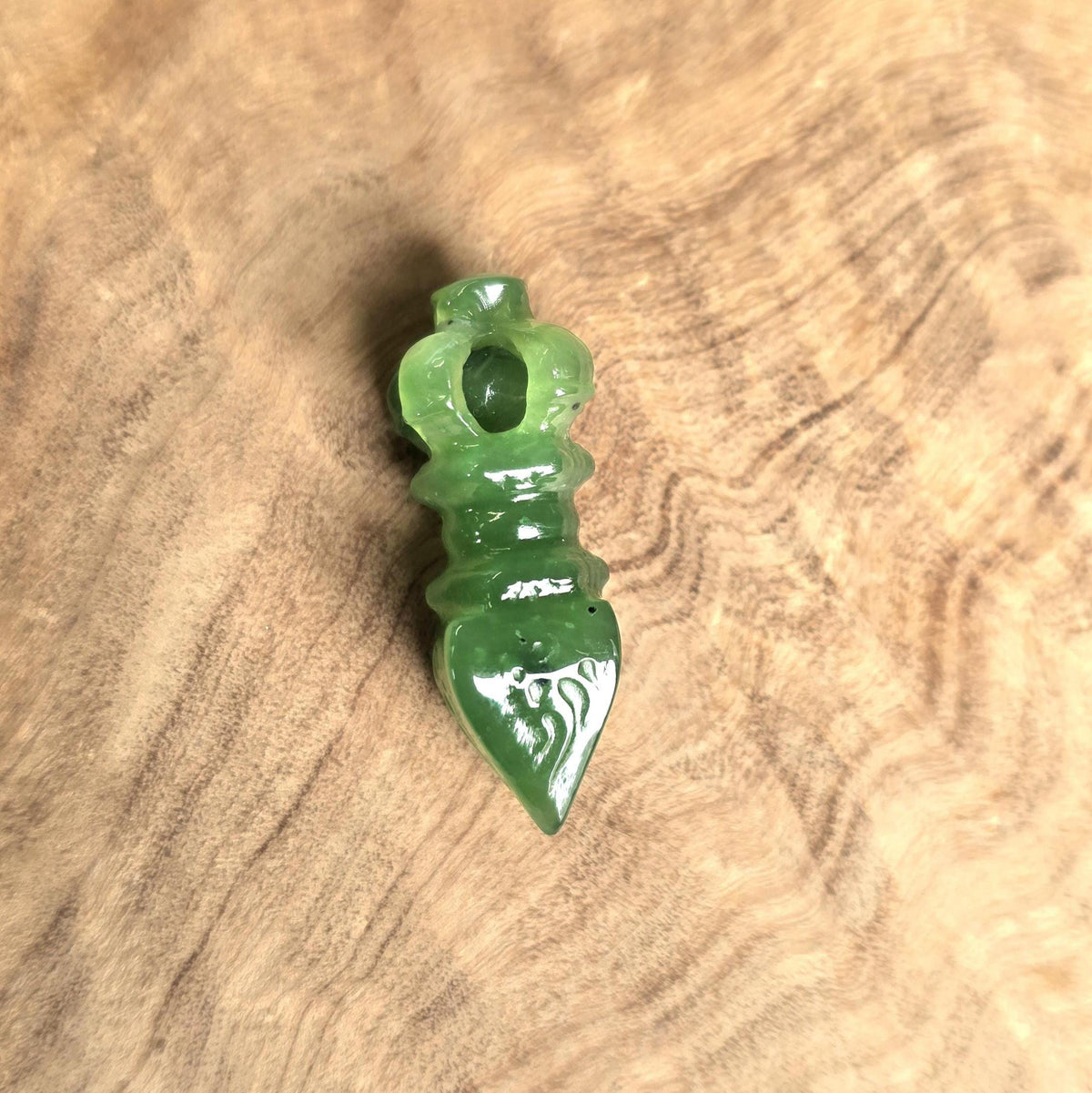 A small carved green stone pendant or charm with a pointed bottom and loop at the top, photographed on a natural wood surface.