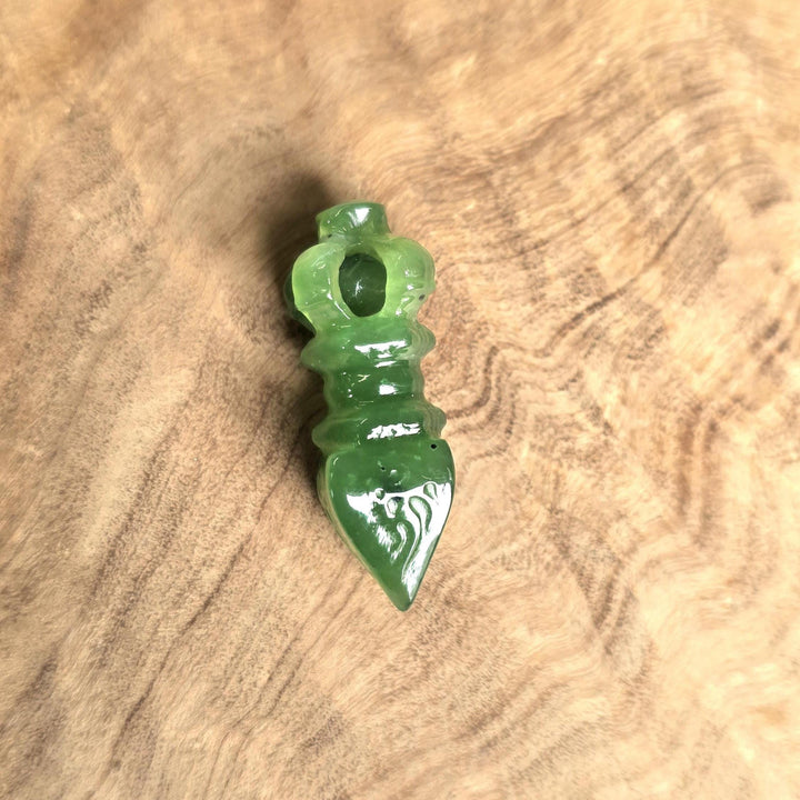 A small carved green stone pendant or charm with a pointed bottom and loop at the top, photographed on a natural wood surface.