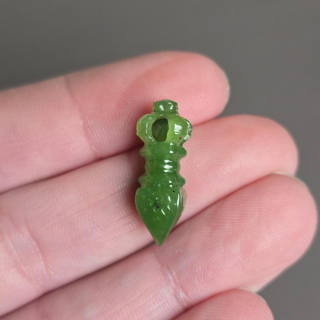 A small carved green jade pendant shaped like a stylized figure, held between fingers to show its size against a neutral gray background.