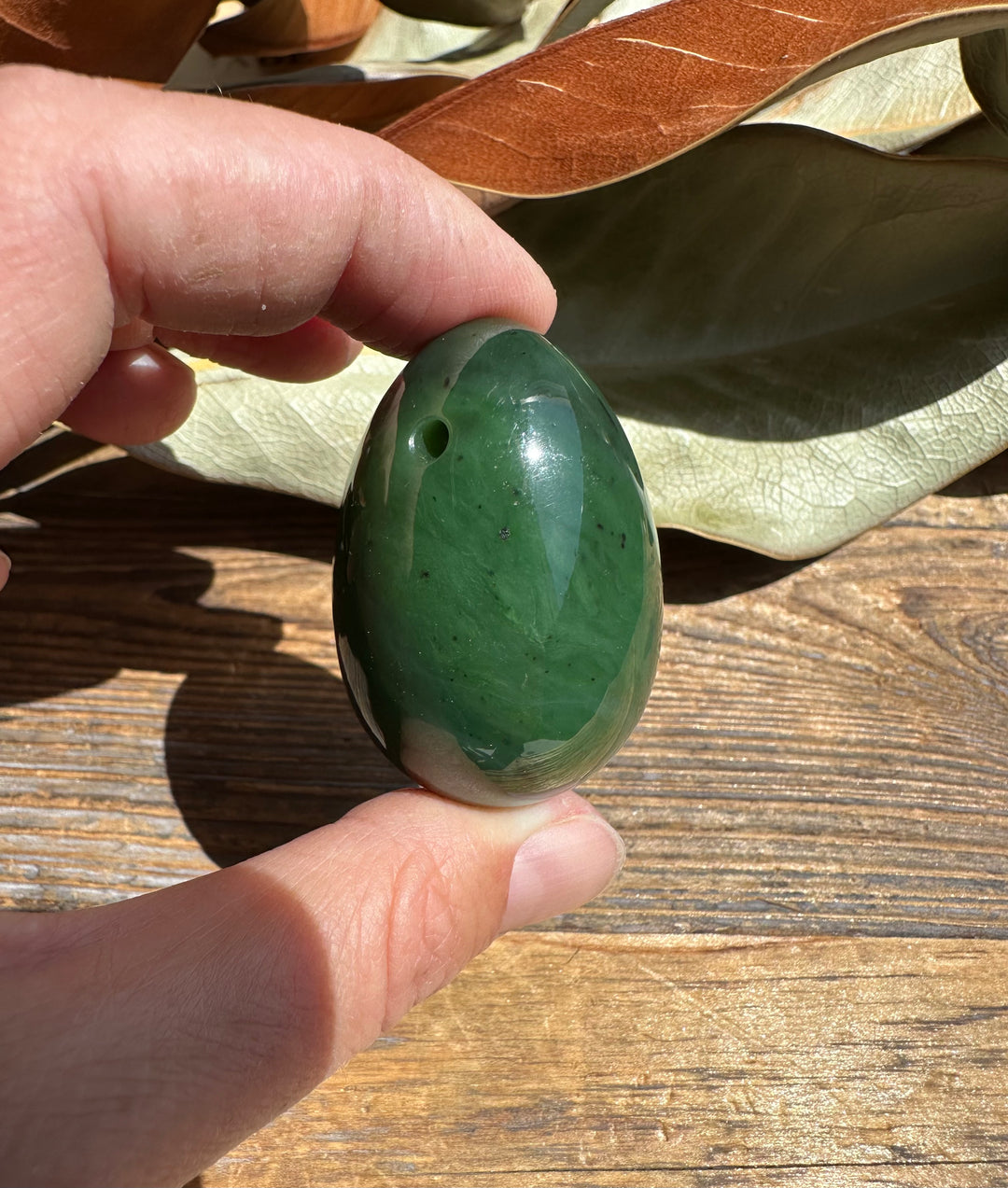 Hand holds a Canadian nephrite yoni egg 1.75 inches, glossy green stone with a drilled hole.