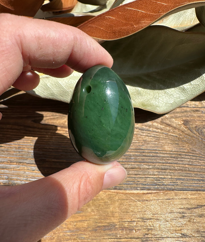 Hand holds a Canadian nephrite yoni egg 1.75 inches, glossy green stone with a drilled hole.