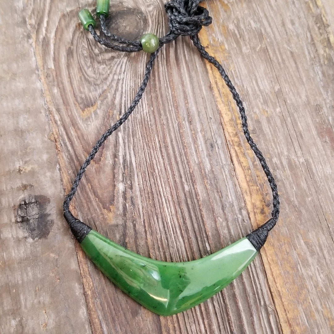 Curved green jade pendant on a black braided cord necklace against a rustic wooden background