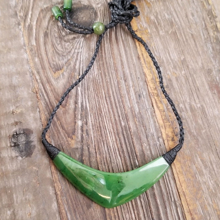 Curved green jade pendant on a black braided cord necklace against a rustic wooden background