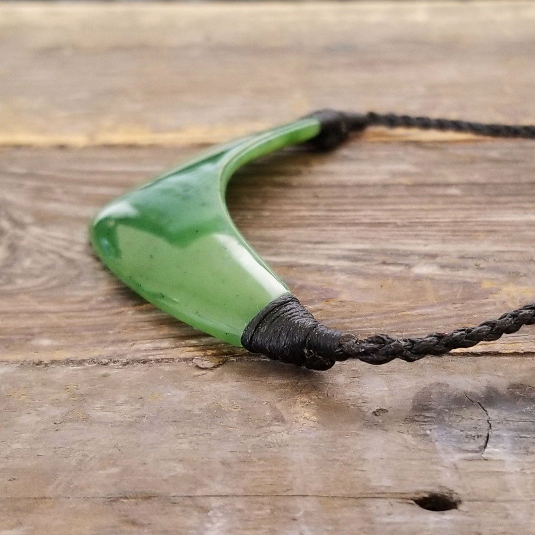 Green jade crescent pendant on a black braided cord resting on a wooden surface