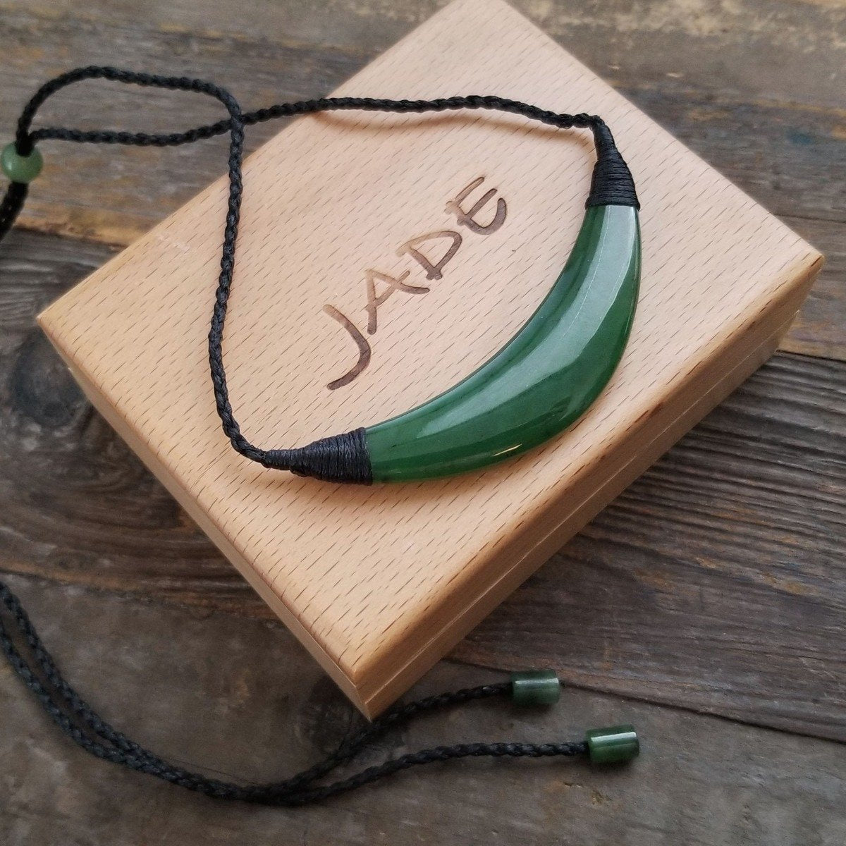 Green curved jade pendant on a black braided cord with green bead accents, displayed on a light wooden box.