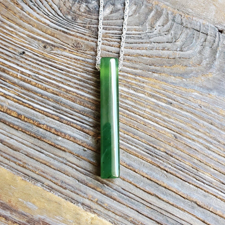 Vertical green jade pendant on a silver chain resting on weathered wood.