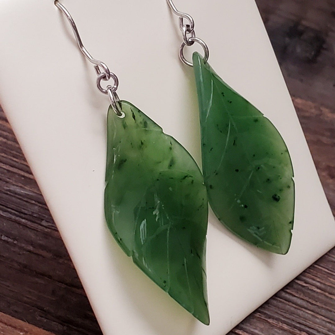 Close-up of jade leaf earrings showing two glossy green leaves with dark flecks and silver hooks.