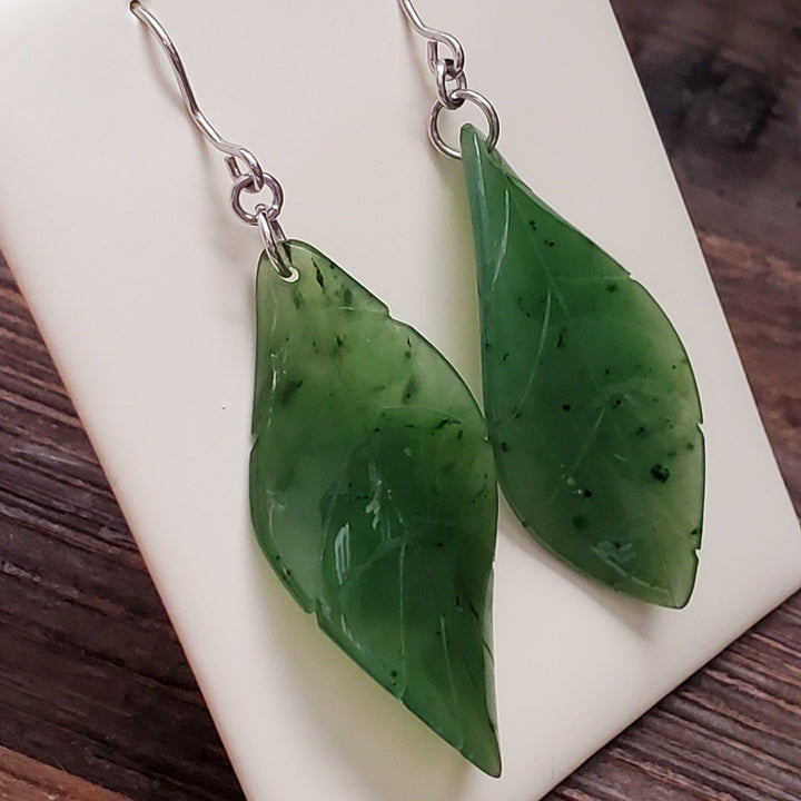 Close-up of jade leaf earrings showing two glossy green leaves with dark flecks and silver hooks.