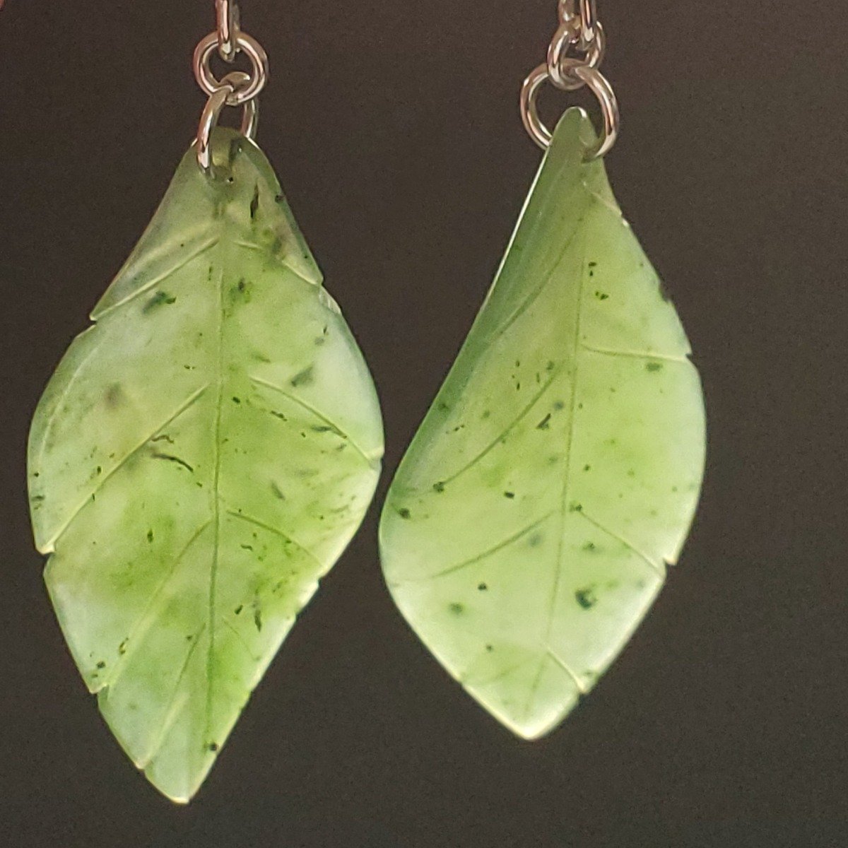 Two pale green jade leaf earrings with silver hooks on a dark background.