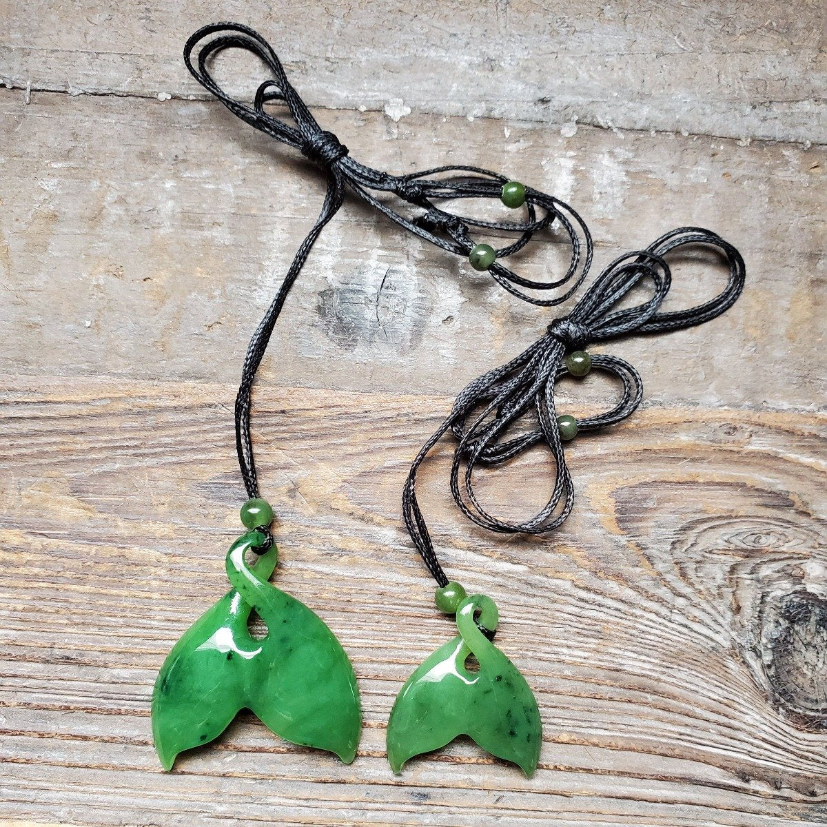 Two green solid whale tail pendants on black cords with green beads on a rustic wooden surface.
