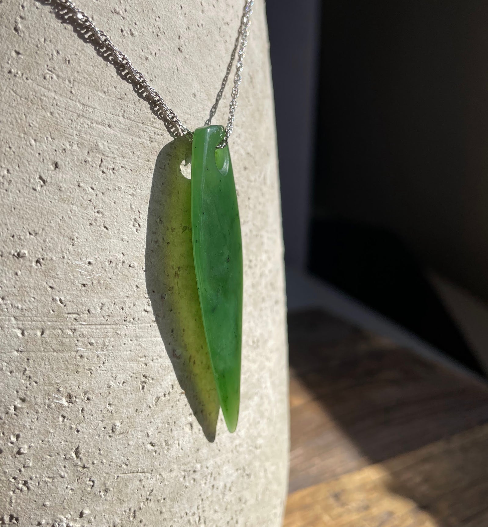 Green jade drop pendant with a top hole on a silver chain against a textured light surface.
