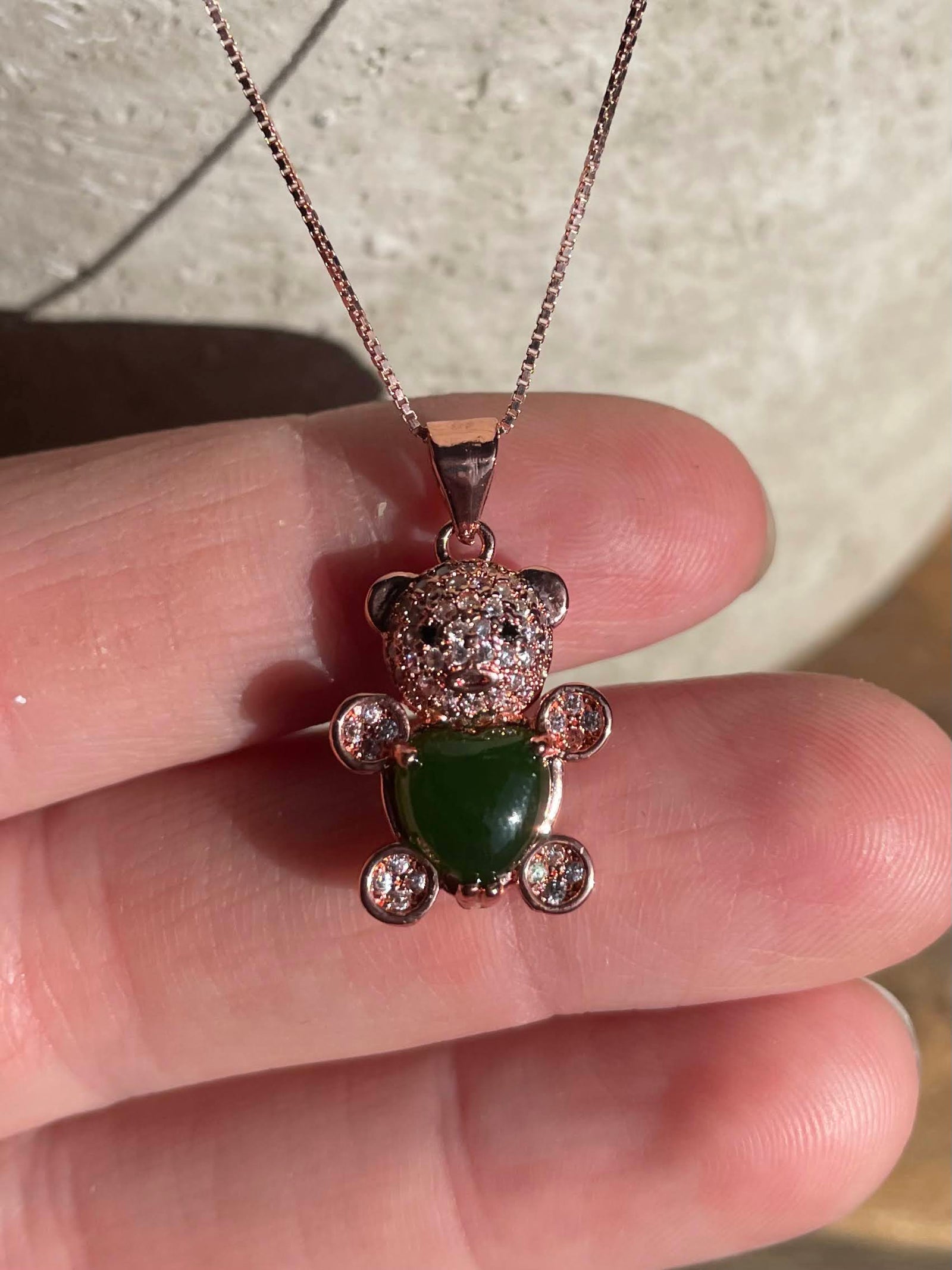 Jade bear pendant with crystal encrusted head and green heart shaped jade body on a rose gold chain held between fingers