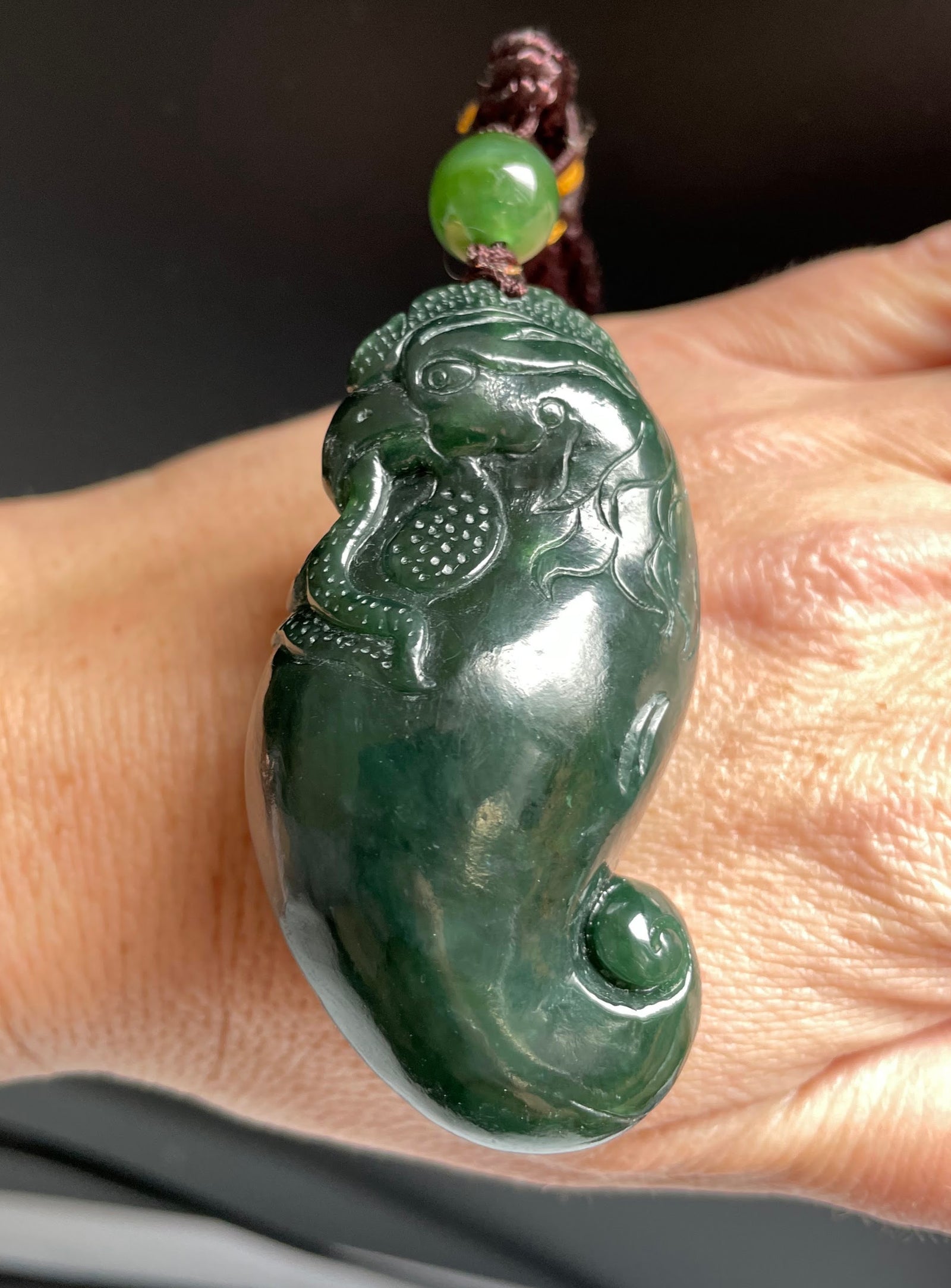 Close up of green jade rooster pendant with bead and braided cord resting on a finger