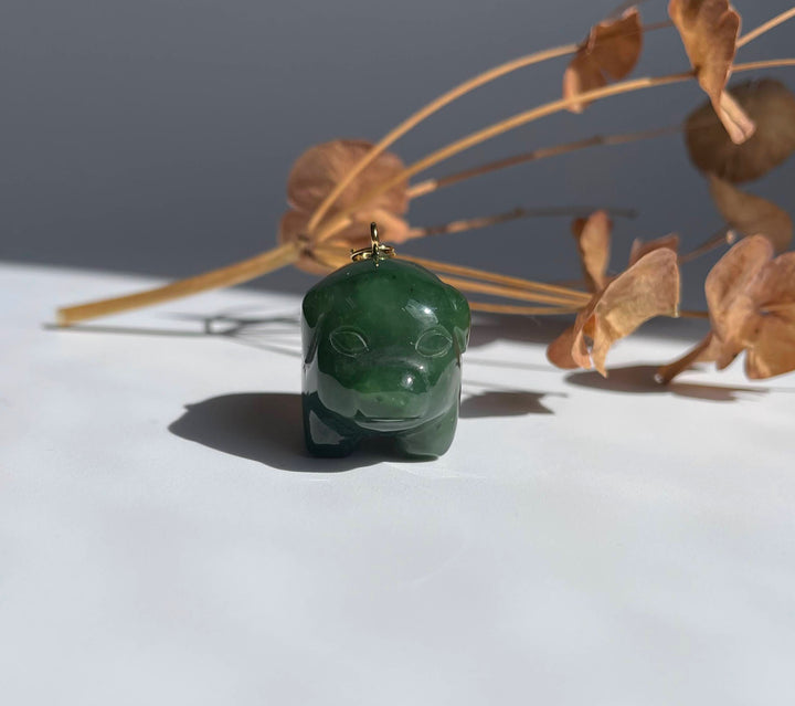 Green jade pig charm with a gold loop sits on a white surface with dried beige plant stems in the background.