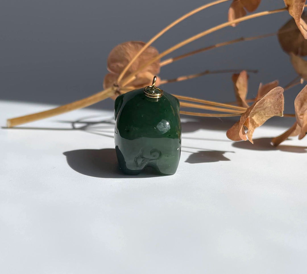 Green jade pig charm with a gold loop on top on a white surface with dried stems in the background.
