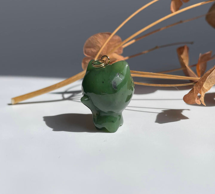 Green jade cartoon cat charm with a small gold loop on top resting on a white surface with dried stems behind.