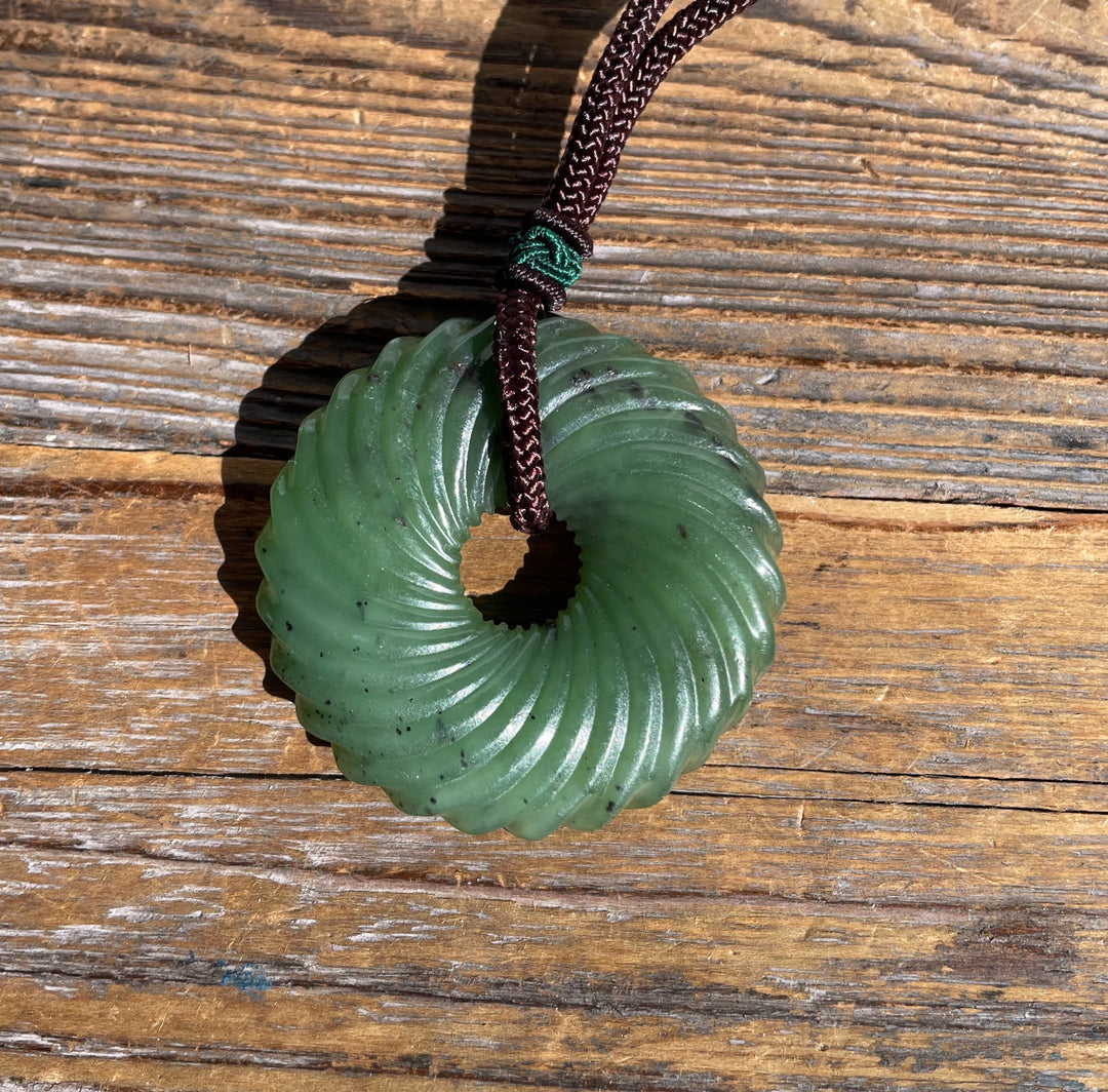 Green jade pendant with circular grooves and central hole, attached to a dark braided cord against a weathered wooden surface.