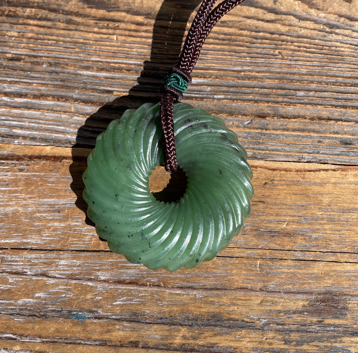Green jade pendant with circular grooves and central hole, attached to a dark braided cord against a weathered wooden surface.