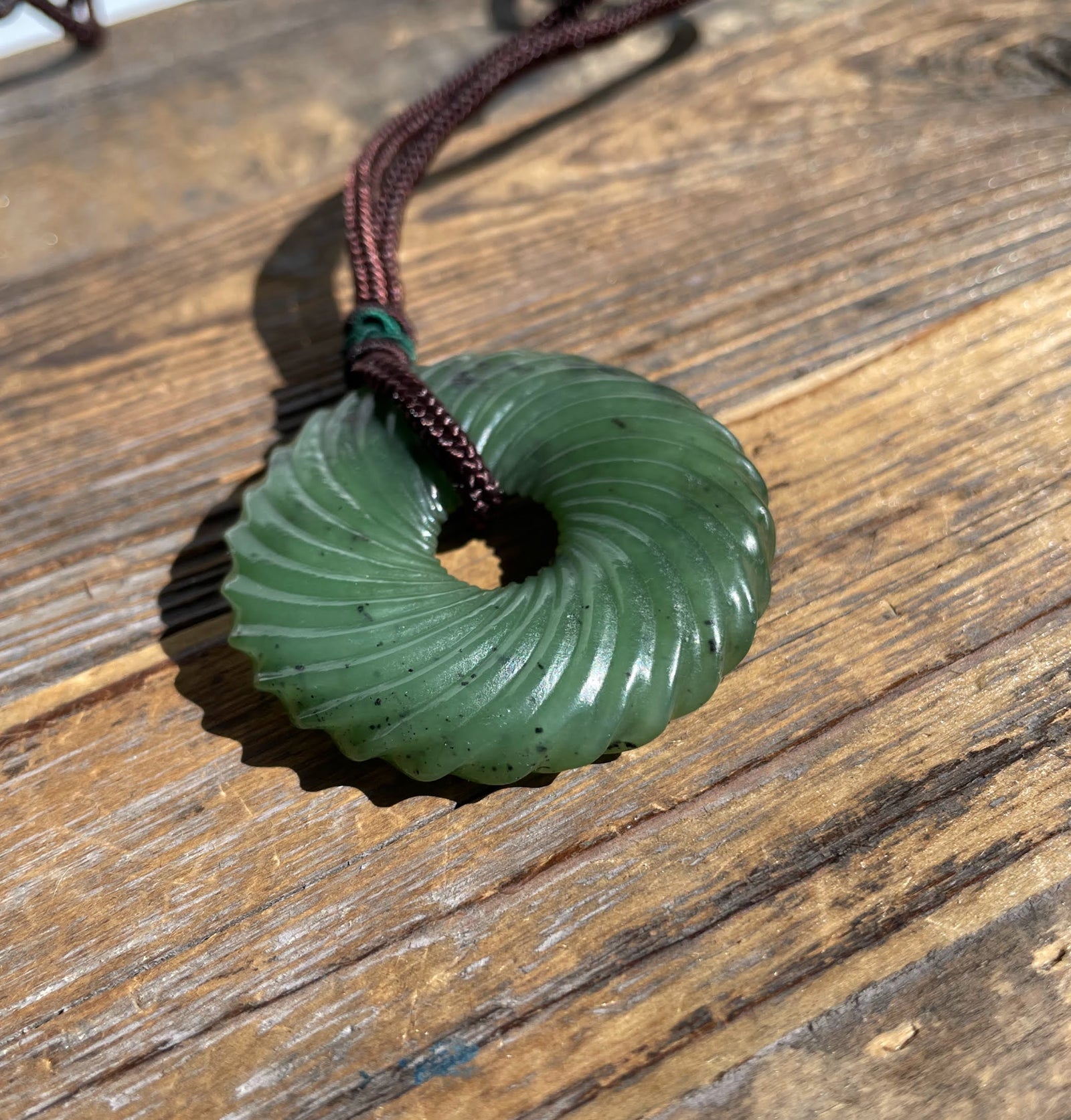 Round green jade pendant with twisted circular ridges and central hole on a brown cord resting on weathered wood.