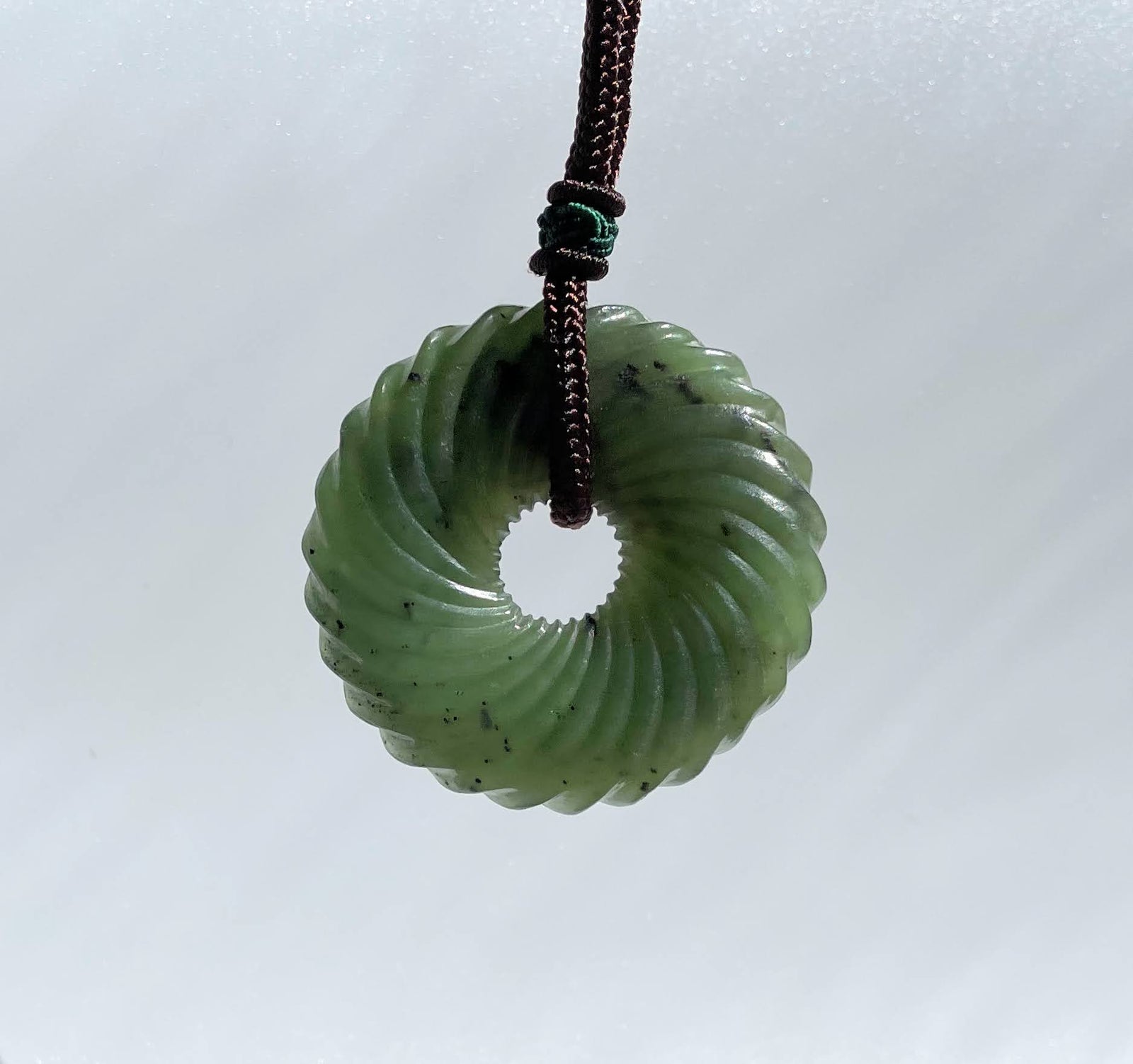 Round twist jade pendant with ribbed donut form and central hole, hanging from a brown cord.