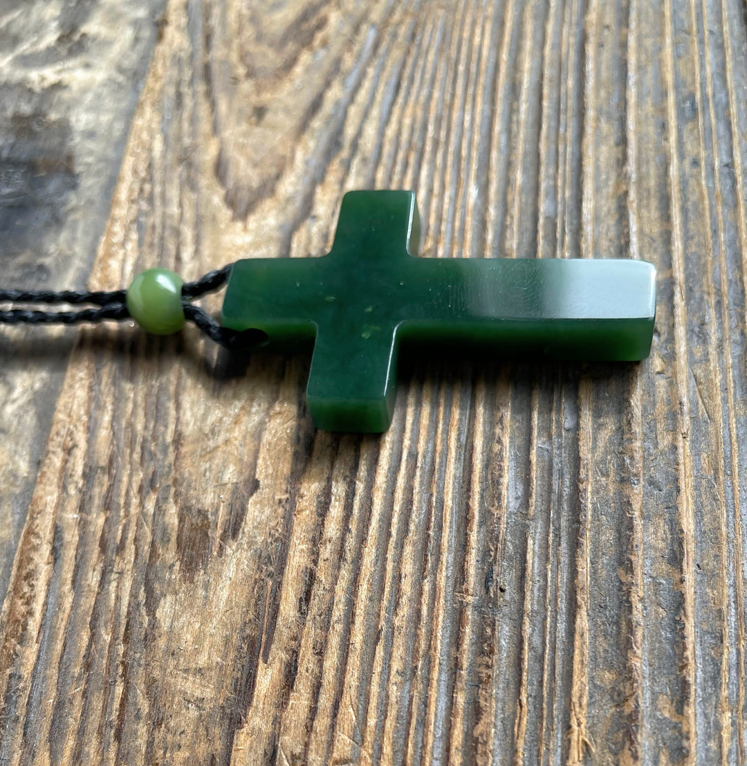 Dark green Canadian jade cross pendant 40mm on a black cord with a green bead on a wooden surface