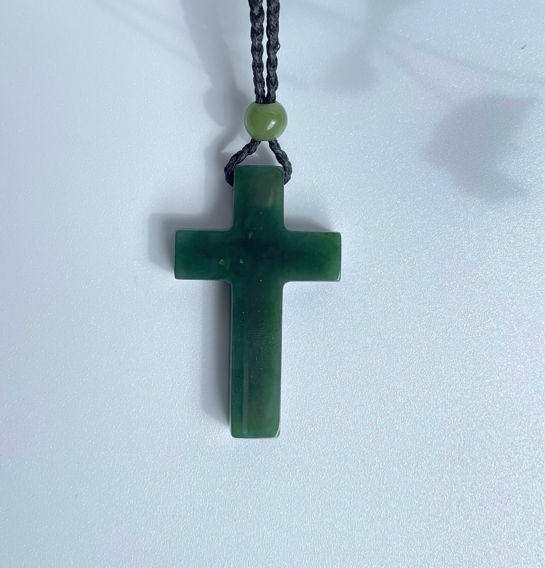 Dark green jade cross pendant with black cord and round bead against a pale background