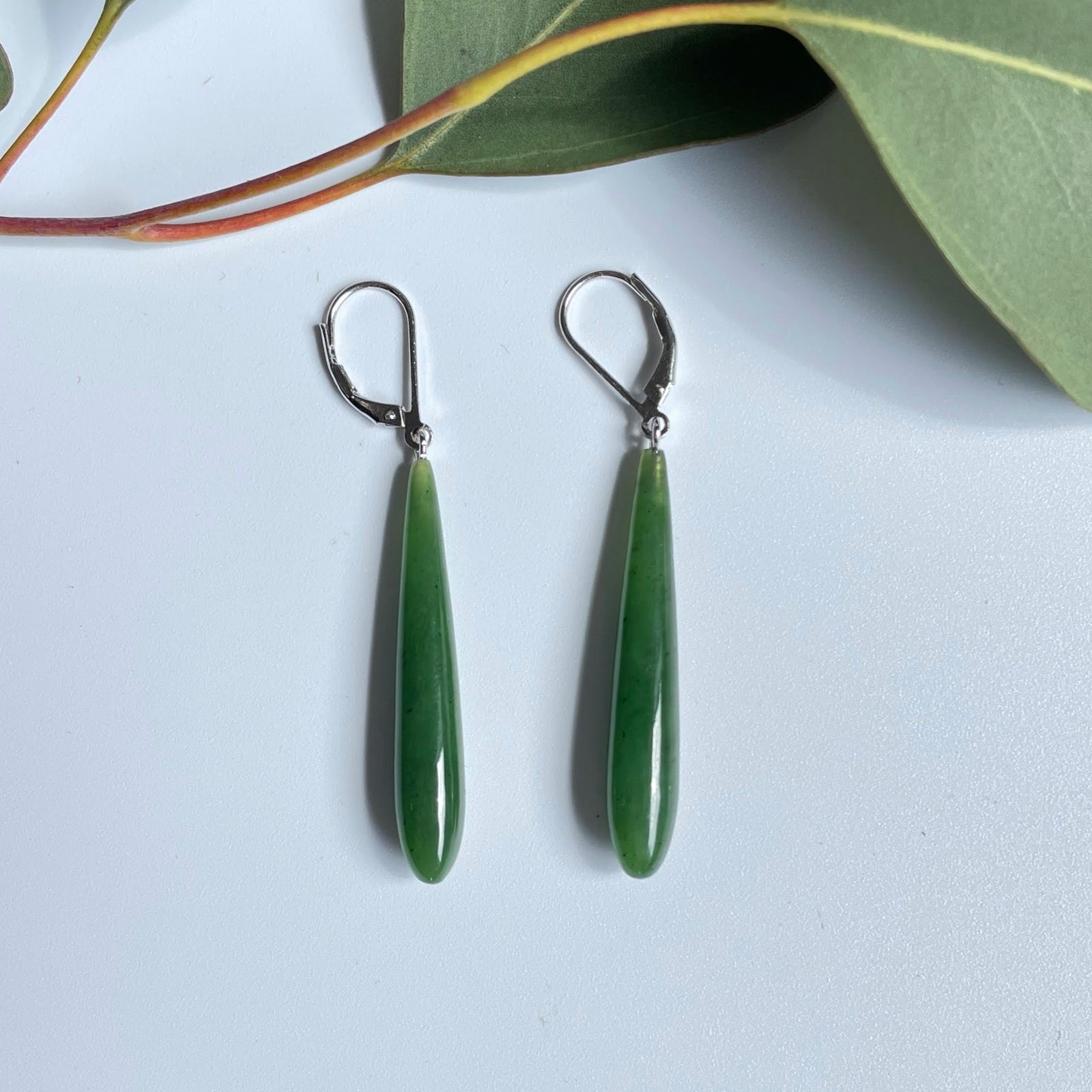 Long jade drop earrings with silver hooks laid on a white surface with green leaves