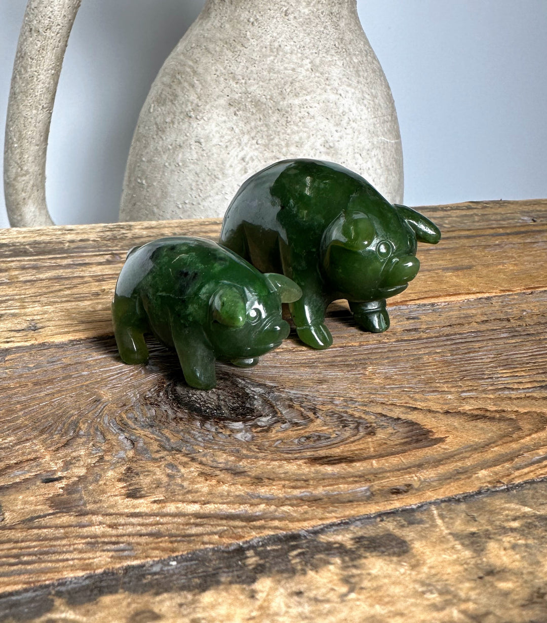 Two green jade pig figurines in different sizes on a weathered wooden surface with a light ceramic jug in the background.