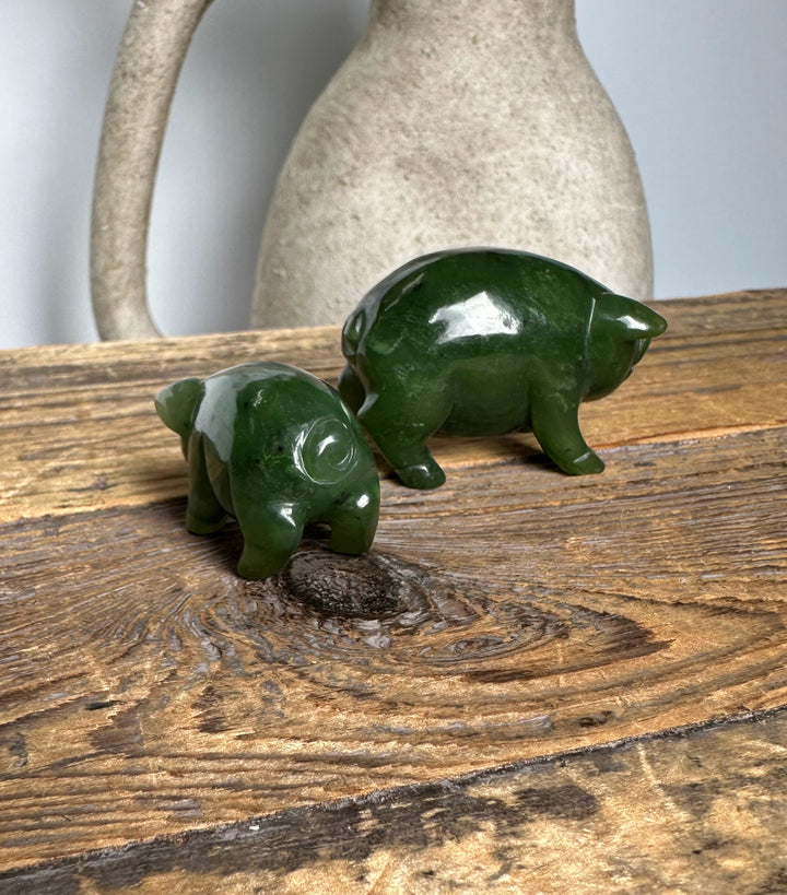 Smaller and larger jade pig statues in rich green on a rustic wood table with a cream ceramic jug behind them.