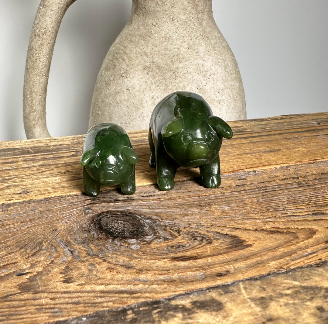 Pair of dark green jade pig figurines on a textured wooden tabletop with a pale ceramic jug in the background.