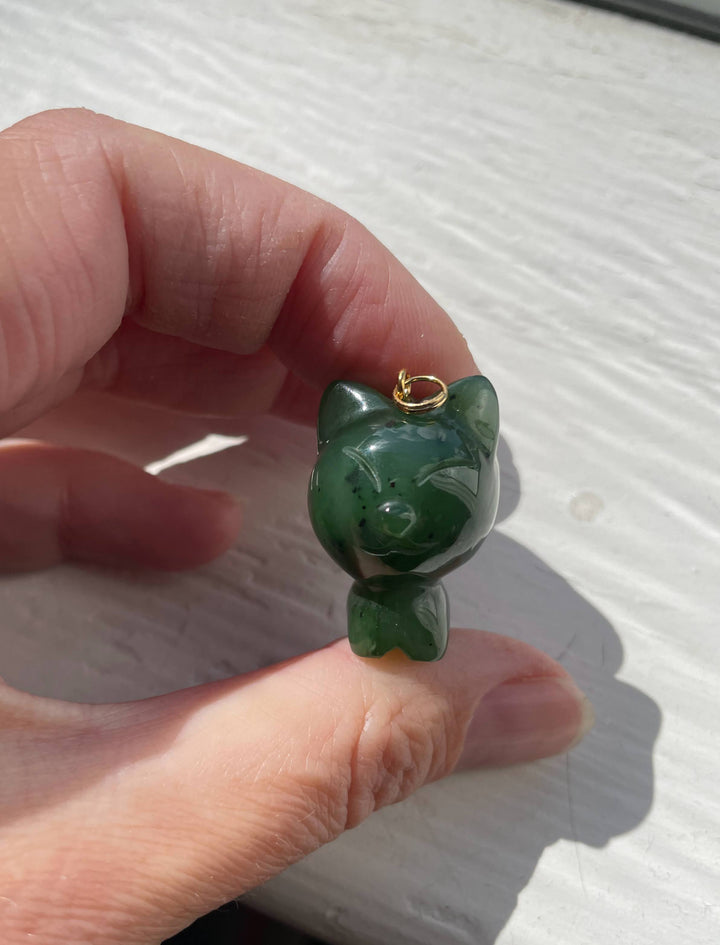 Cartoon cat charm carved from green jade held between fingers outdoors in bright sunlight.