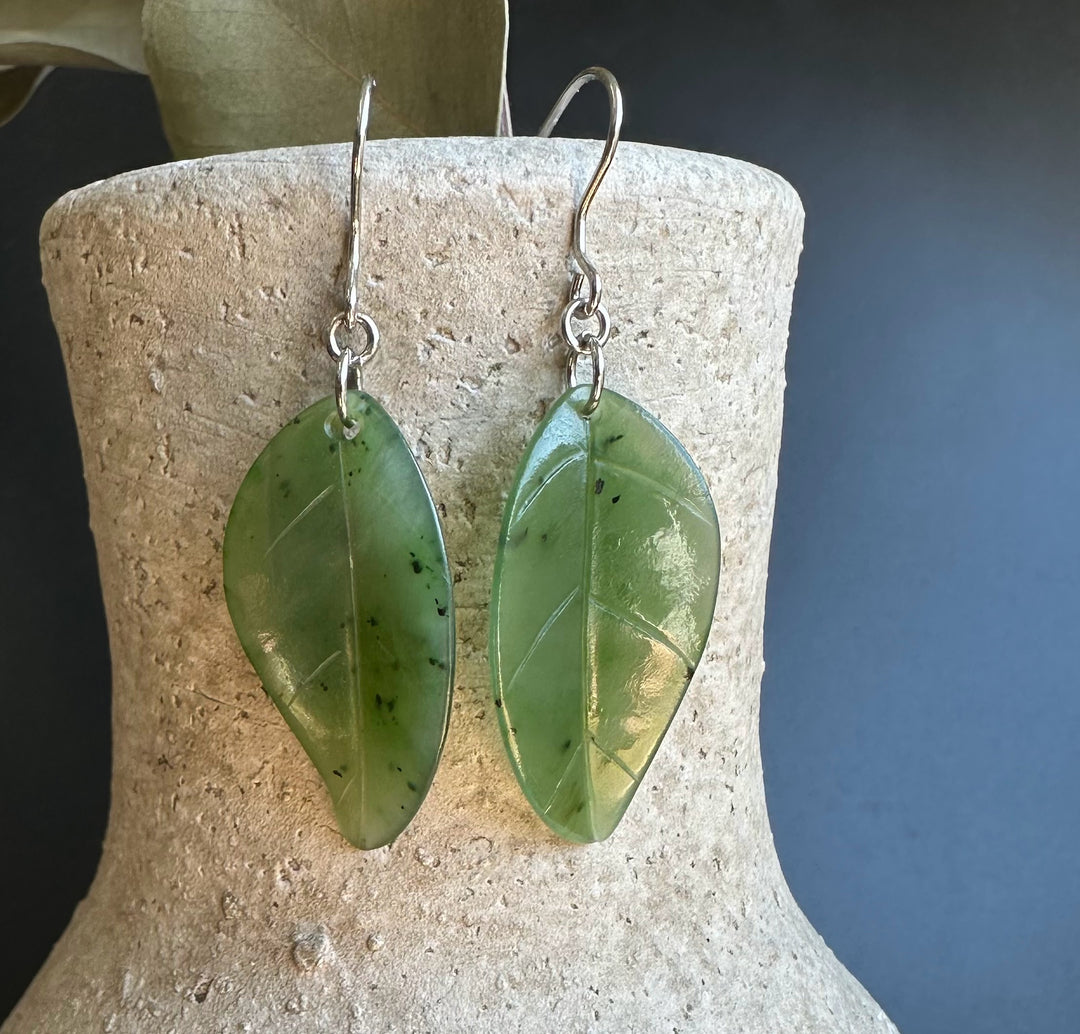 Leaf shaped jade earrings in pale green with silver hooks displayed on a textured beige stand