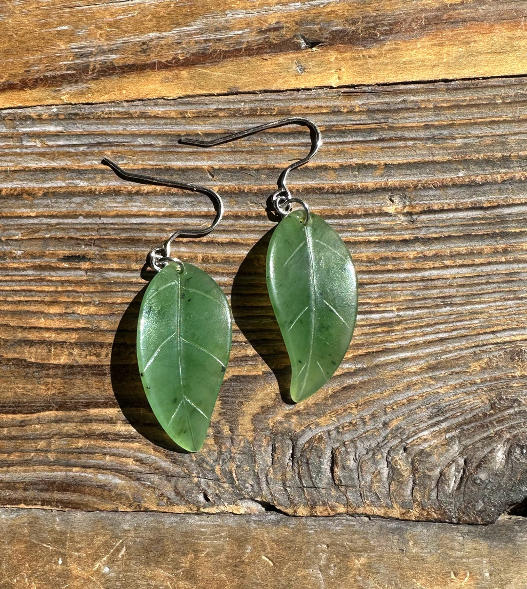 Two jade leaf earrings with silver hooks on a rustic wooden surface